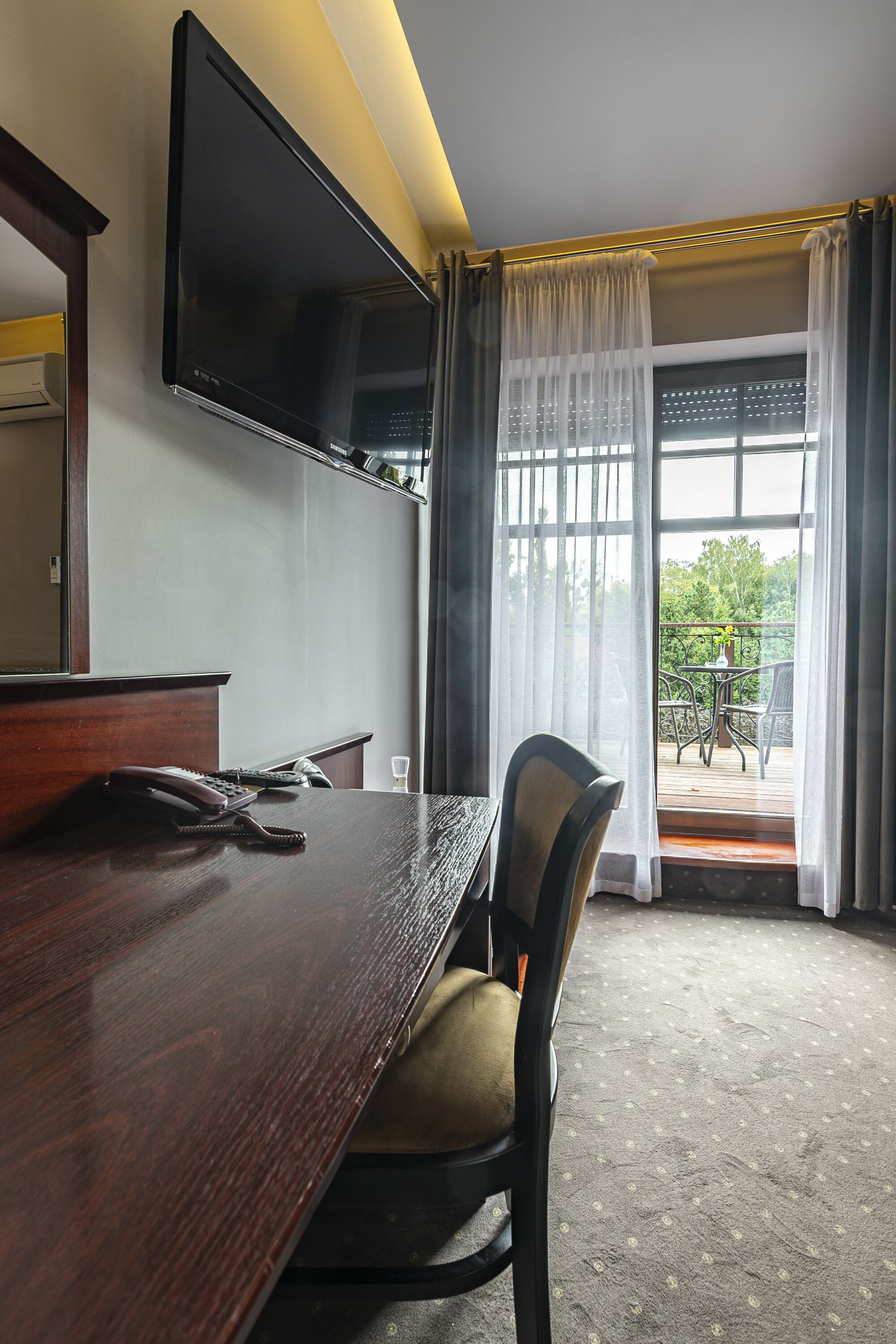 A hotel room with a wooden desk, a flat-screen TV mounted on the wall, a telephone, and a chair. There is a balcony with a table and chairs, and a view of greenery outside.