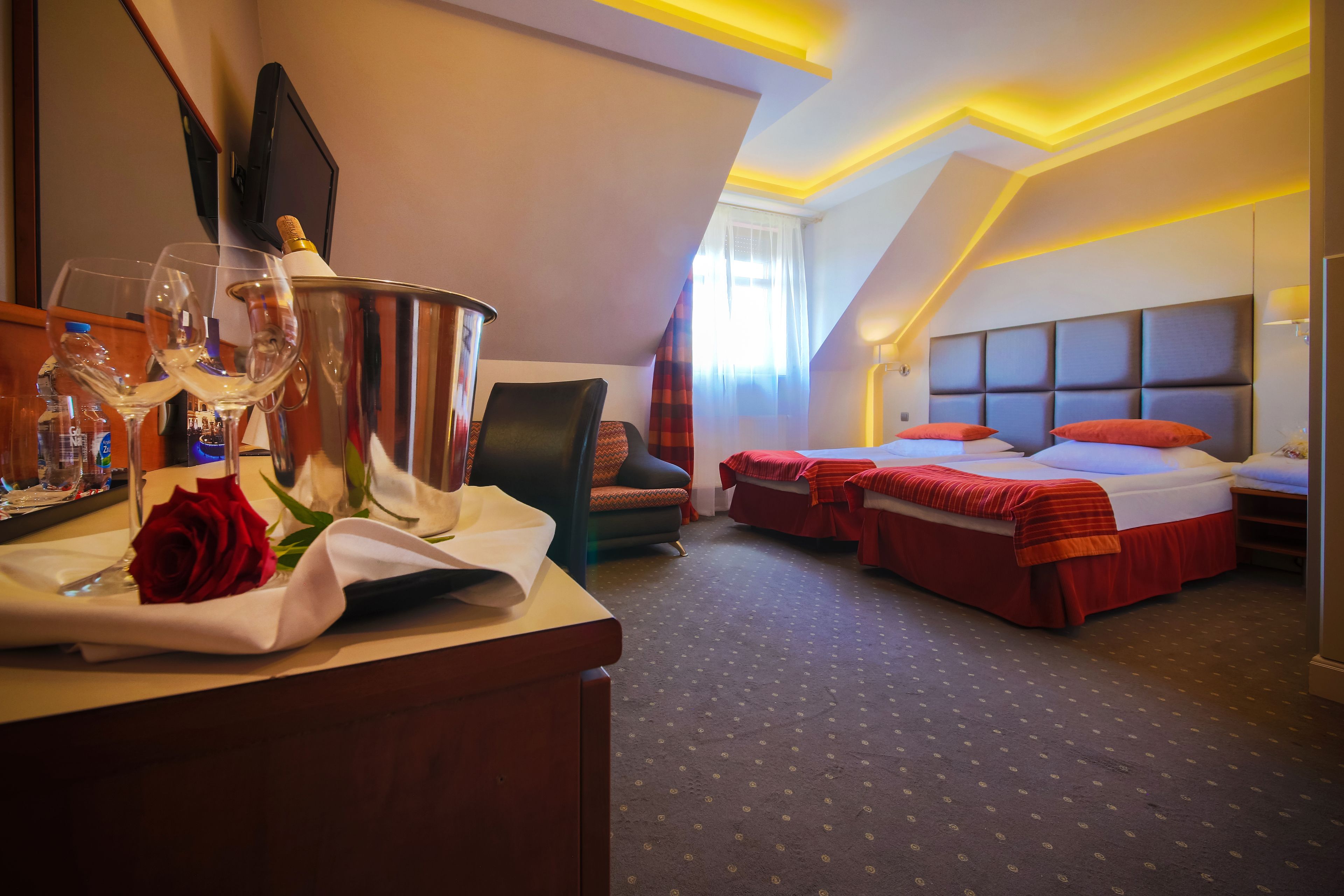 A hotel room with a bed, a chair, a table with a bottle and glasses, and a red rose.