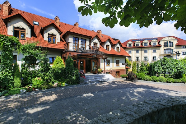 A hotel with a red-tiled roof, a central entrance with steps, and a garden area with various plants and a statue.