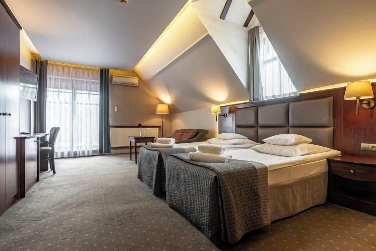 Ein Hotelzimmer mit einem großen Bett, einem Schreibtisch und einem Stuhl. Es gibt ein Fenster mit Vorhängen, eine Lampe und ein an der Wand befestigtes Fernsehgerät.