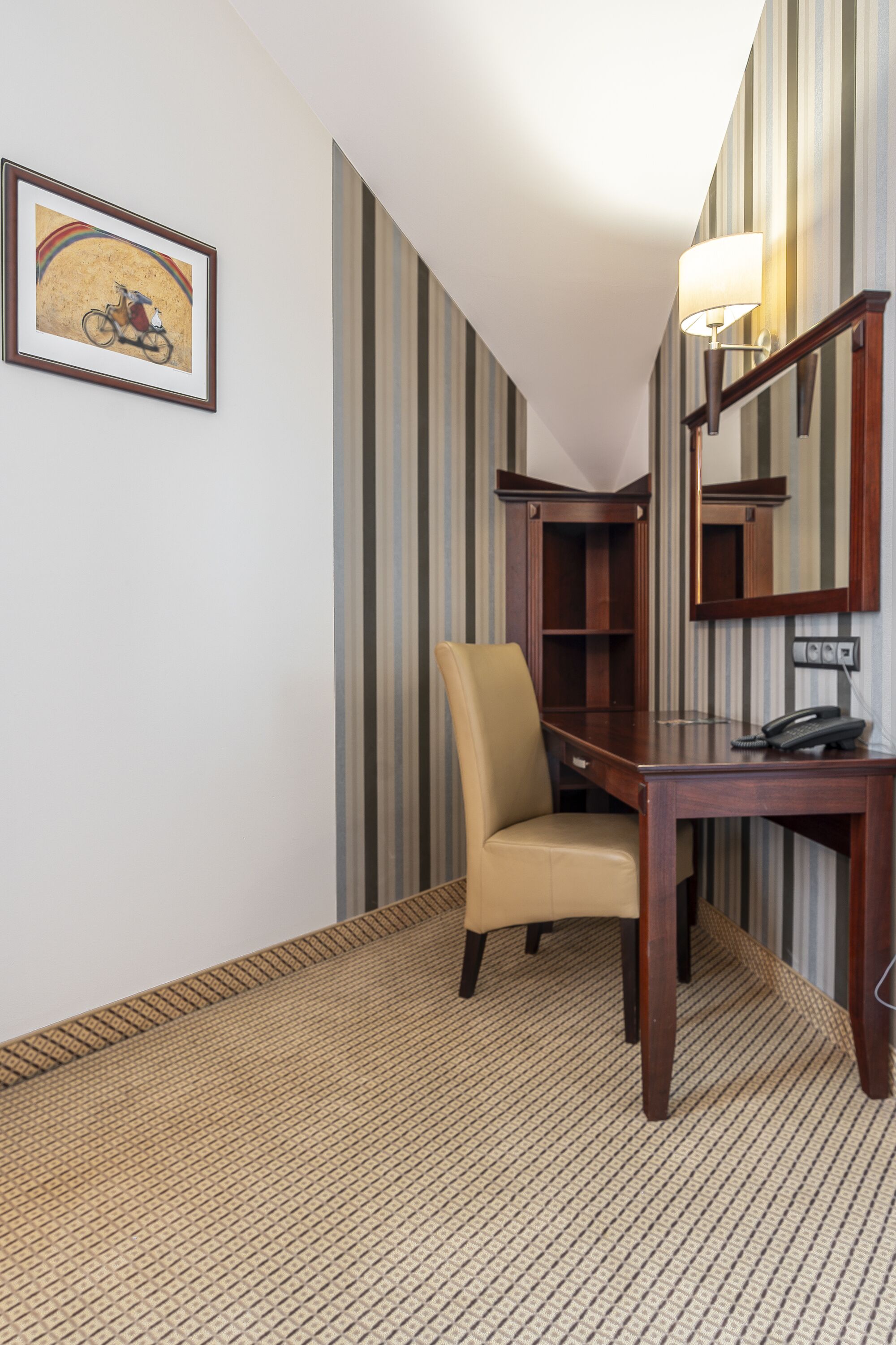 A hotel room with a wooden desk, a chair, a telephone, a lamp, a bookshelf, and a framed picture on the wall.