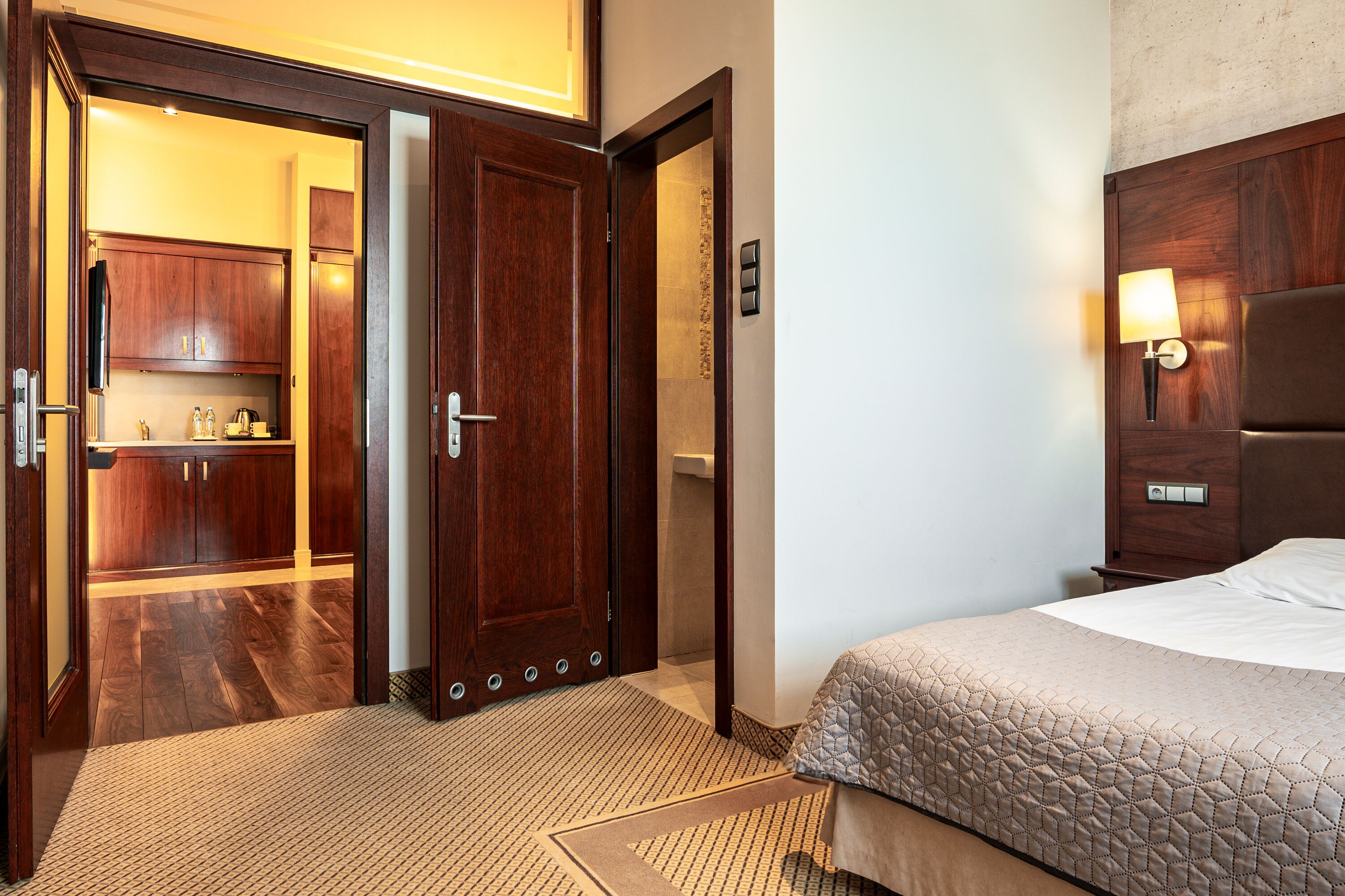A hotel room with a wooden door leading to a kitchen, a bed with a quilted cover, and a wall-mounted light fixture.