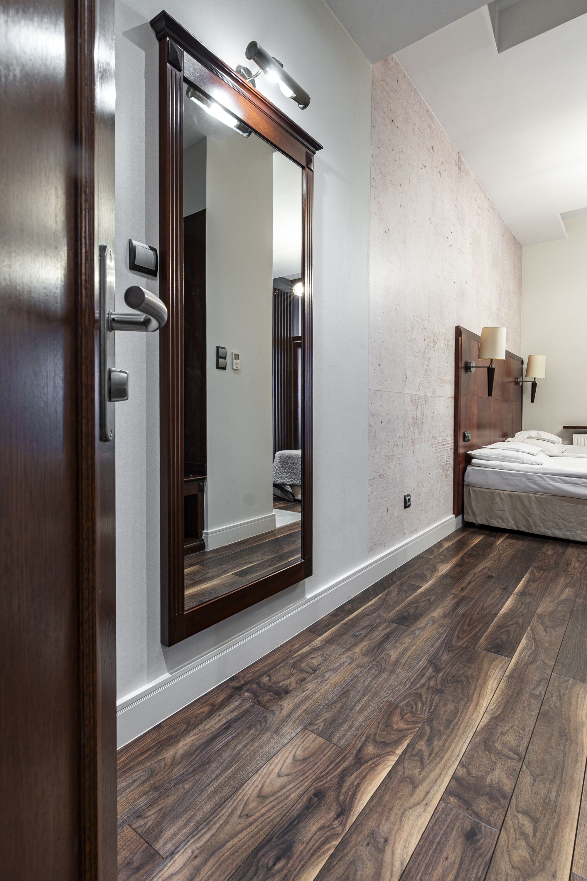 A bedroom with a large mirror, wooden flooring, a bed, and a nightstand.