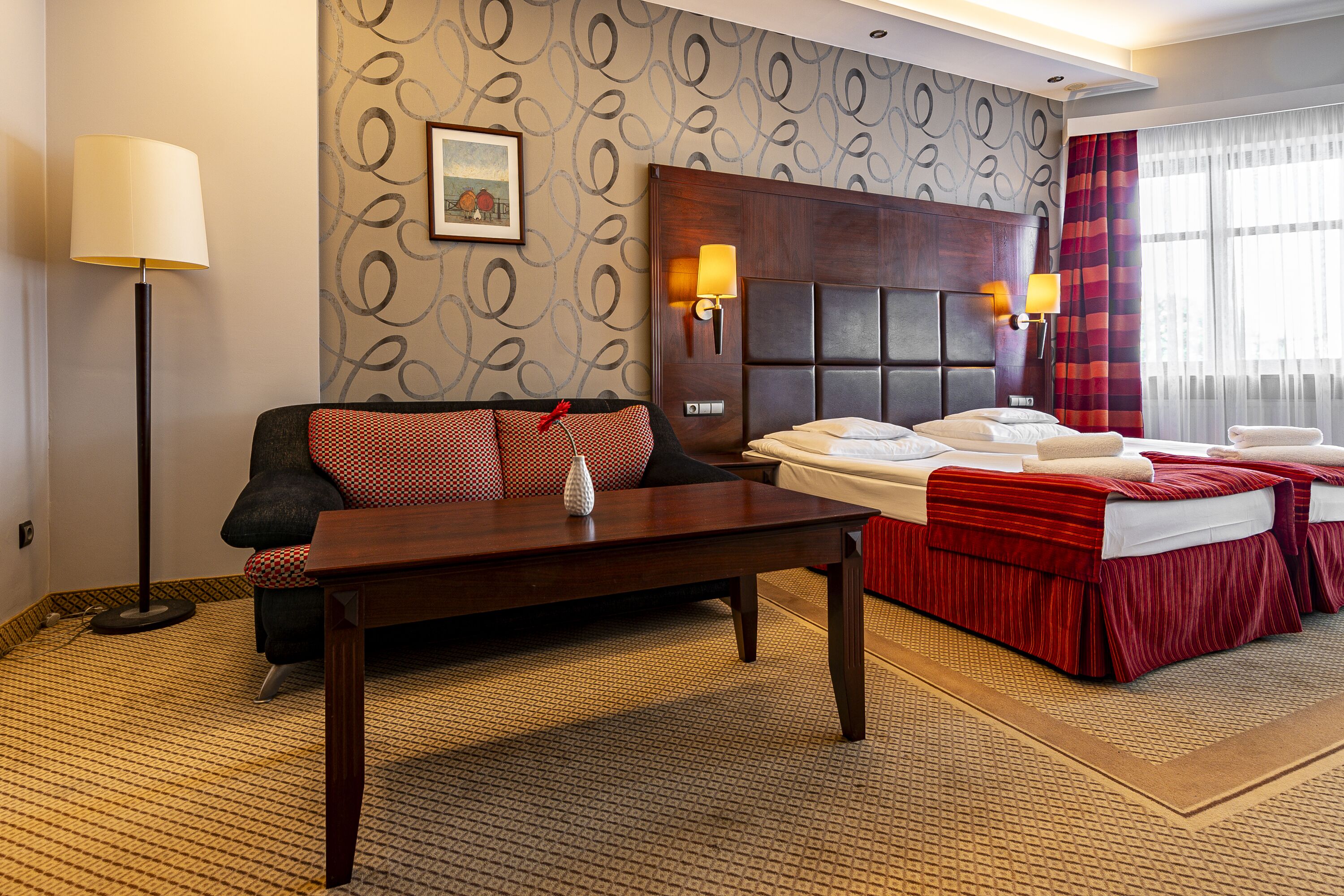 A hotel room with a sofa, a bed with red bedding, a wooden headboard, two wall-mounted lamps, and a patterned carpet.