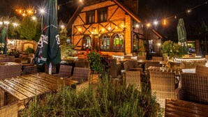 An outdoor seating area with wicker furniture and umbrellas, a brick building with a timber-framed structure, and string lights in the background.