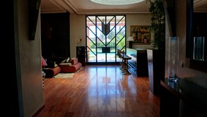 Ein großzügiger Wohnbereich mit Holzboden, einem roten Sofa, einem Fernseher und Blick auf einen Swimmingpool durch eine geometrisch gestaltete Glasschiebetür.