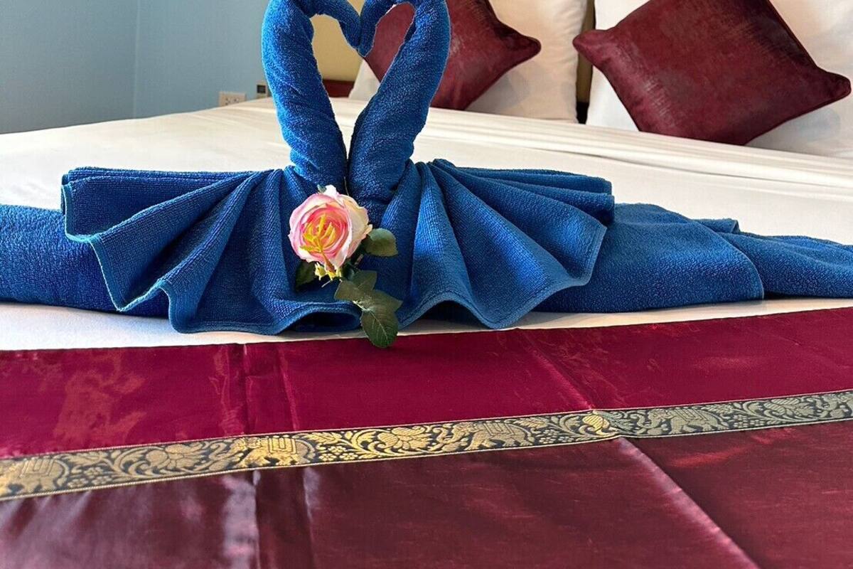 A neatly made bed with a blue towel arranged in a heart shape, a pink flower, and red pillows.