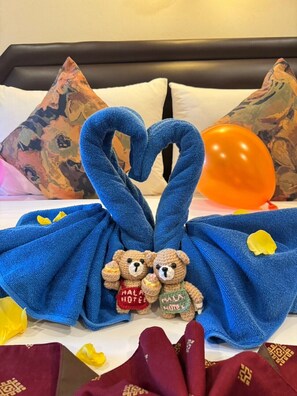 Una cama con una toalla azul dispuesta en forma de corazón, dos osos de peluche y un globo naranja.