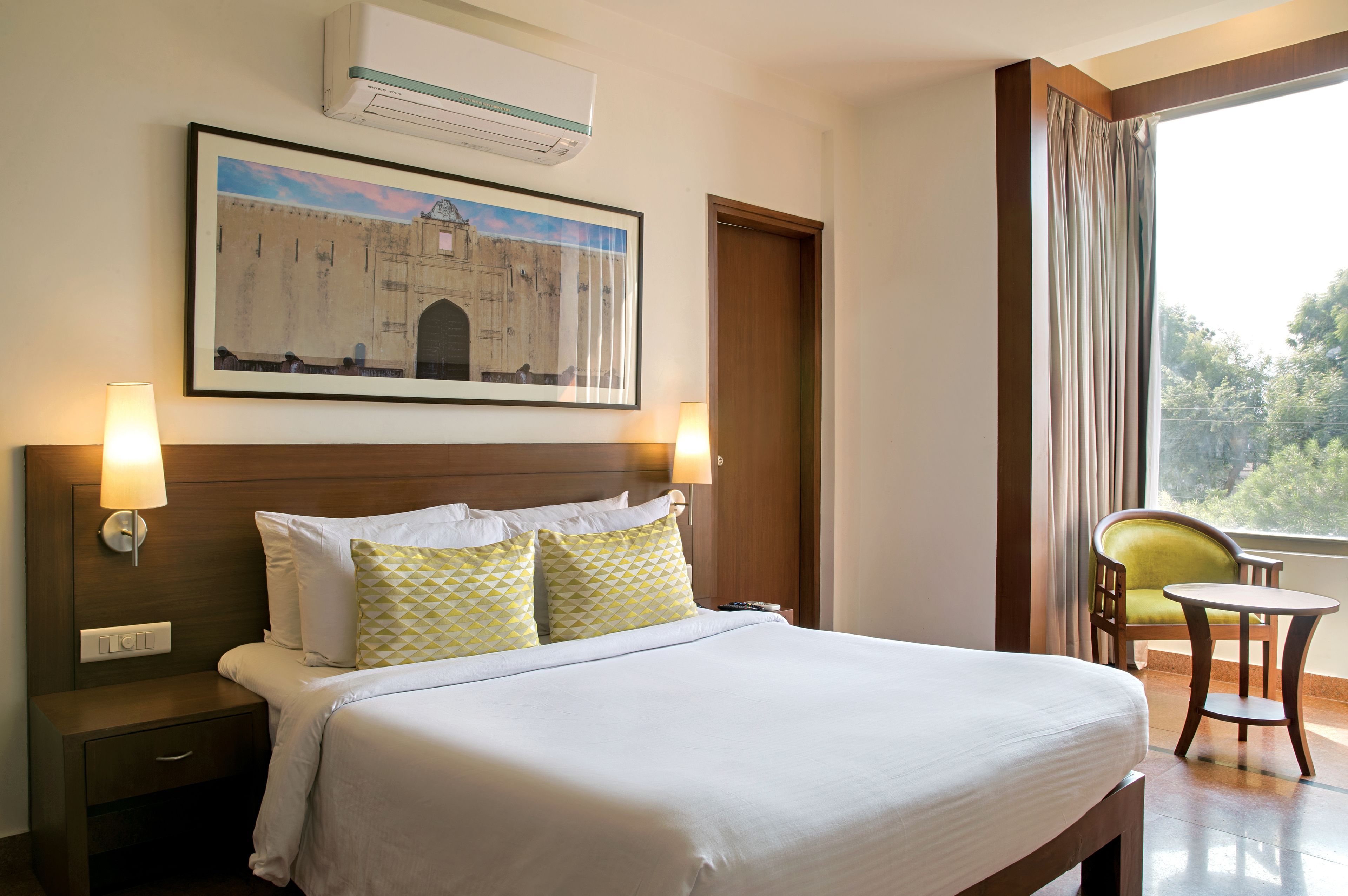 A hotel room with a bed, bedside lamps, a framed picture on the wall, a window with curtains, and a chair by the window.
