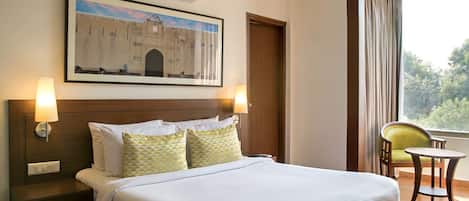 Une chambre d’hôtel avec un lit, des lampes de chevet, un tableau encadré au mur, une fenêtre avec des rideaux et une chaise près de la fenêtre.