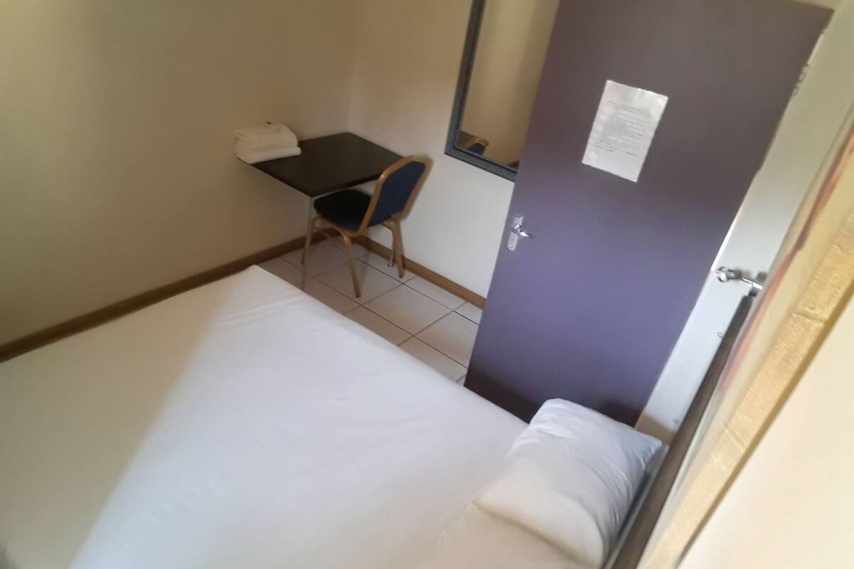 A small, single-bed room with a desk and chair, a door with a notice, and a tiled floor.