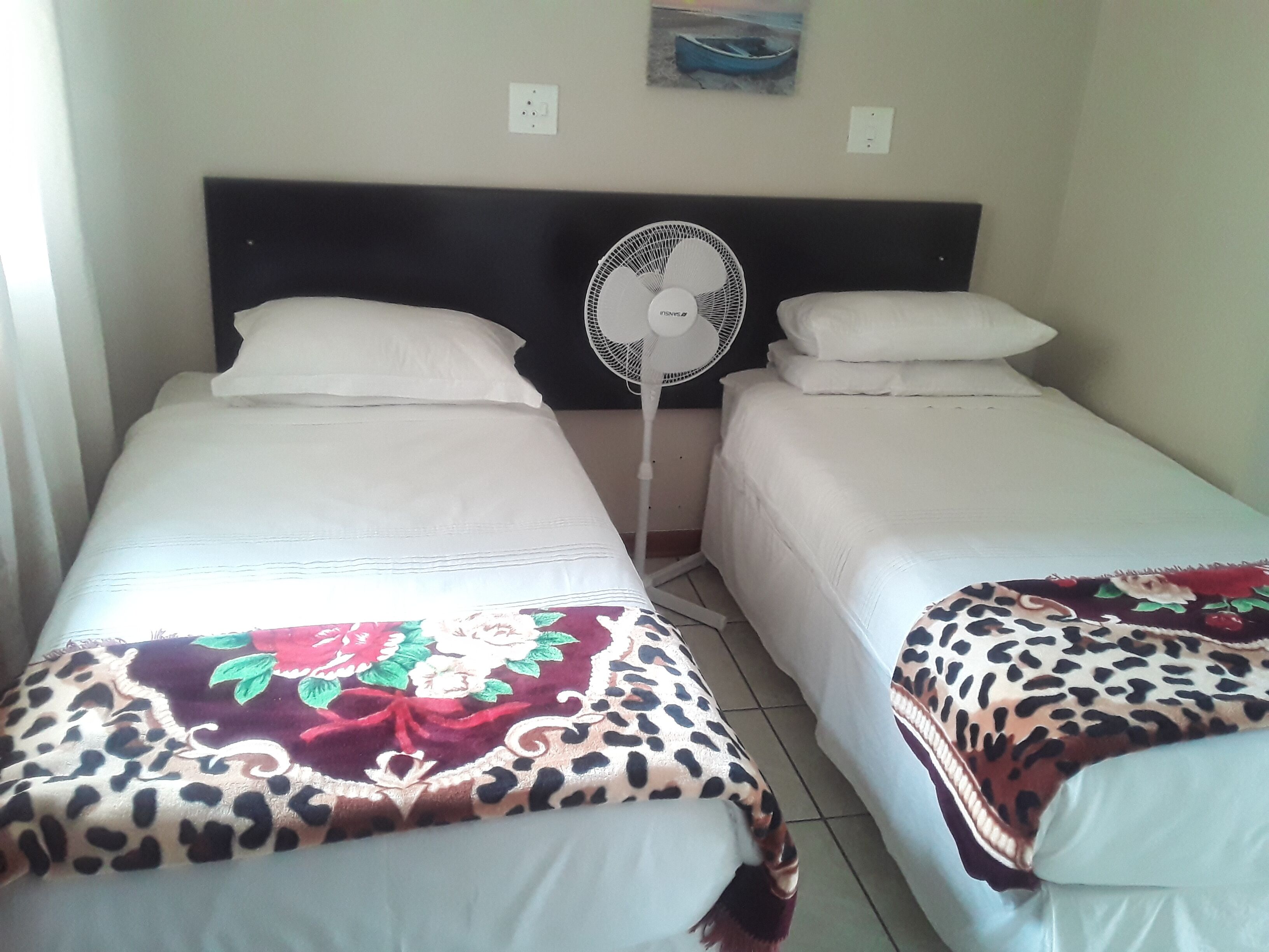 Une chambre avec deux lits simples, chacun recouvert d’une couette blanche et d’une couverture à motifs léopard. Un ventilateur sur pied est placé entre les lits, et un tableau est accroché au mur au-dessus du lit situé à droite.