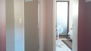 Un pasillo con paredes blancas y suelo de baldosas que conduce a una habitación con un ventilador y una puerta.