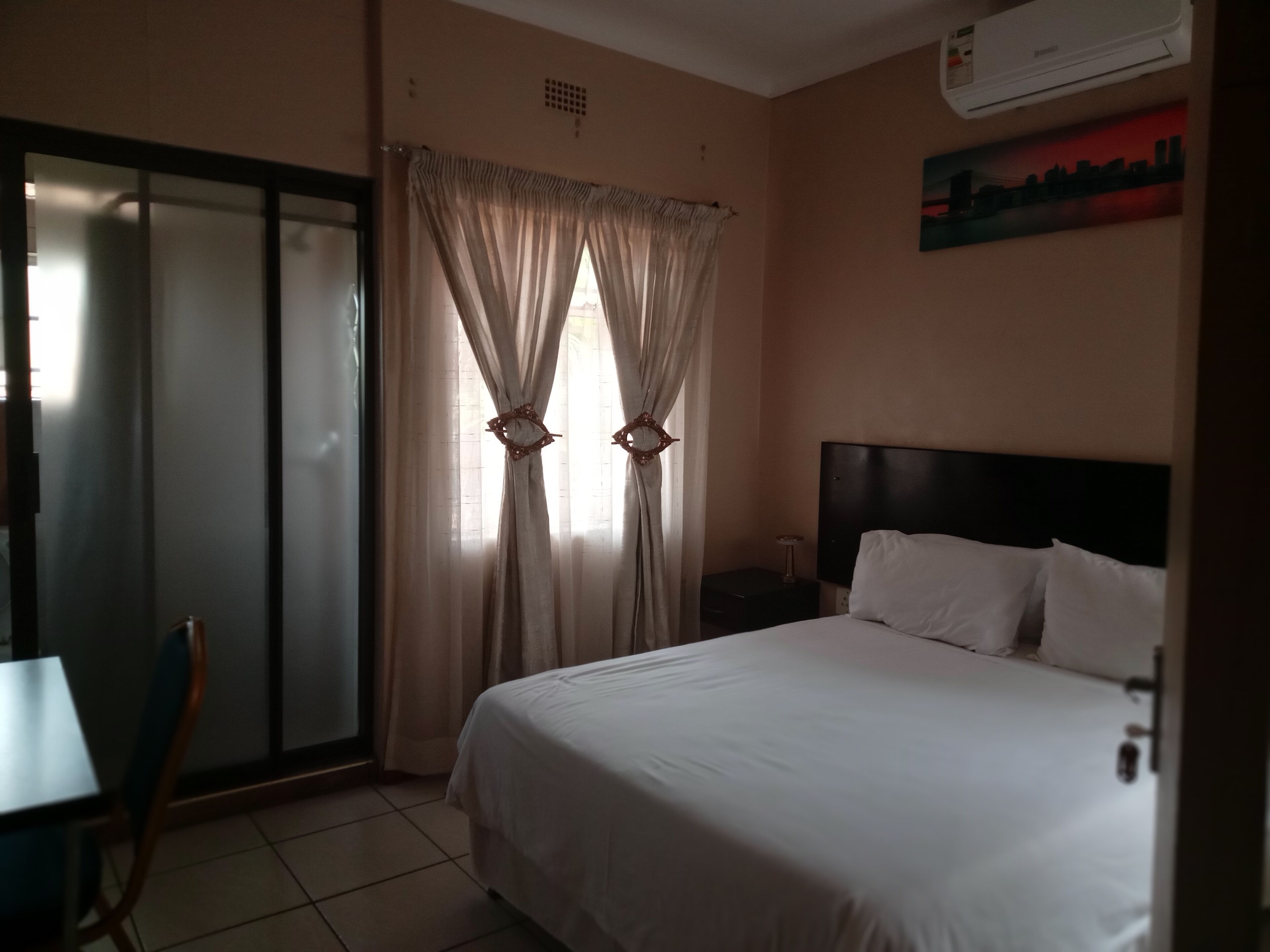 A hotel room with a bed, a desk, and a chair. There is a window with curtains and an air conditioning unit on the wall.