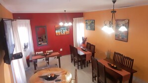 Un área de comedor con una mesa redonda, mesas rectangulares y sillas de madera. Hay dos ventiladores de techo y un cuadro en la pared.
