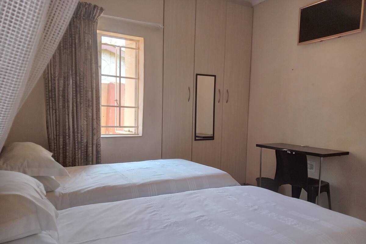 A bedroom with two beds, a window with curtains, a mirror, and a desk.