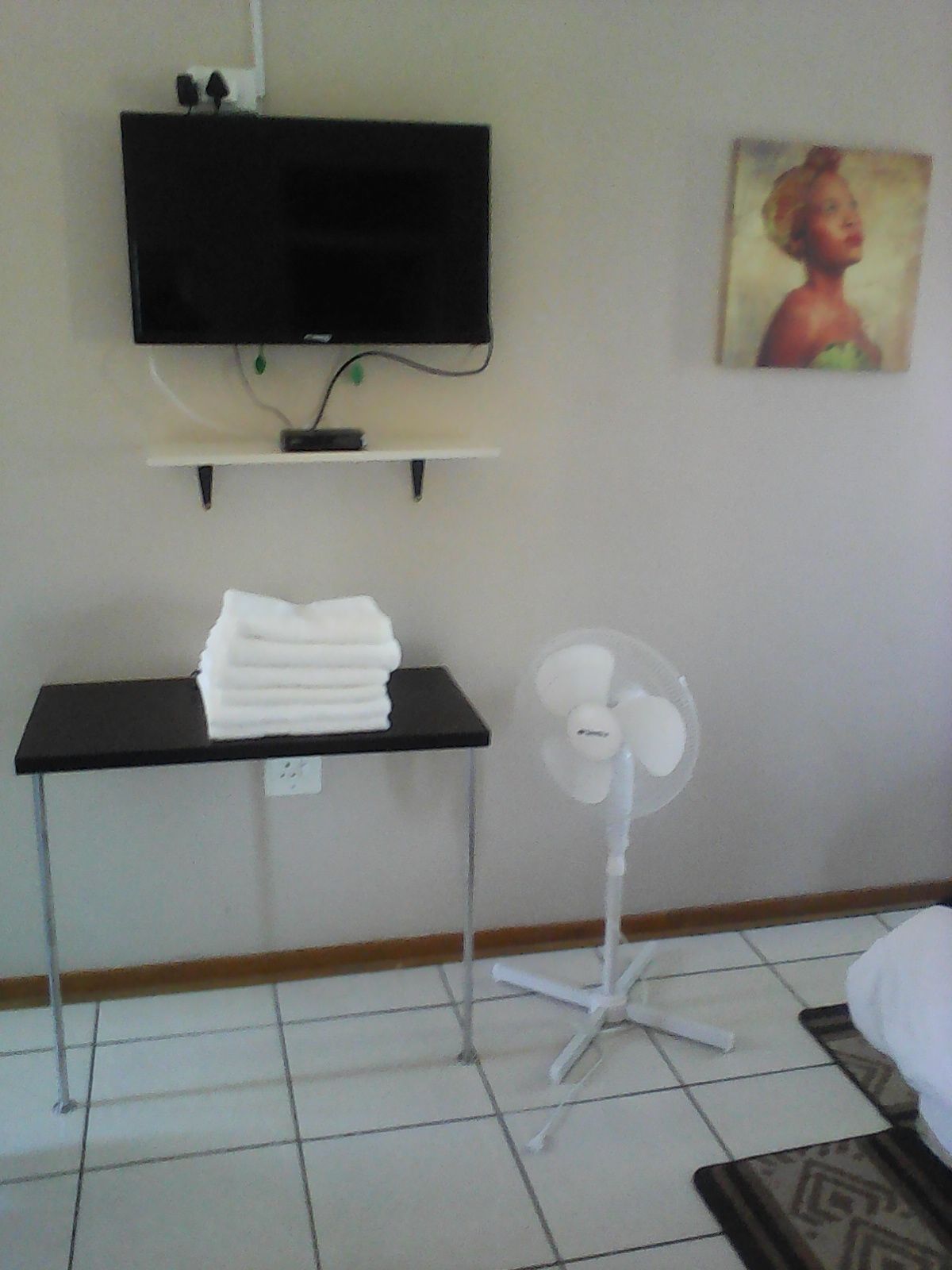 A room with a flat-screen TV mounted on the wall, a table with a stack of white towels, and a standing fan.