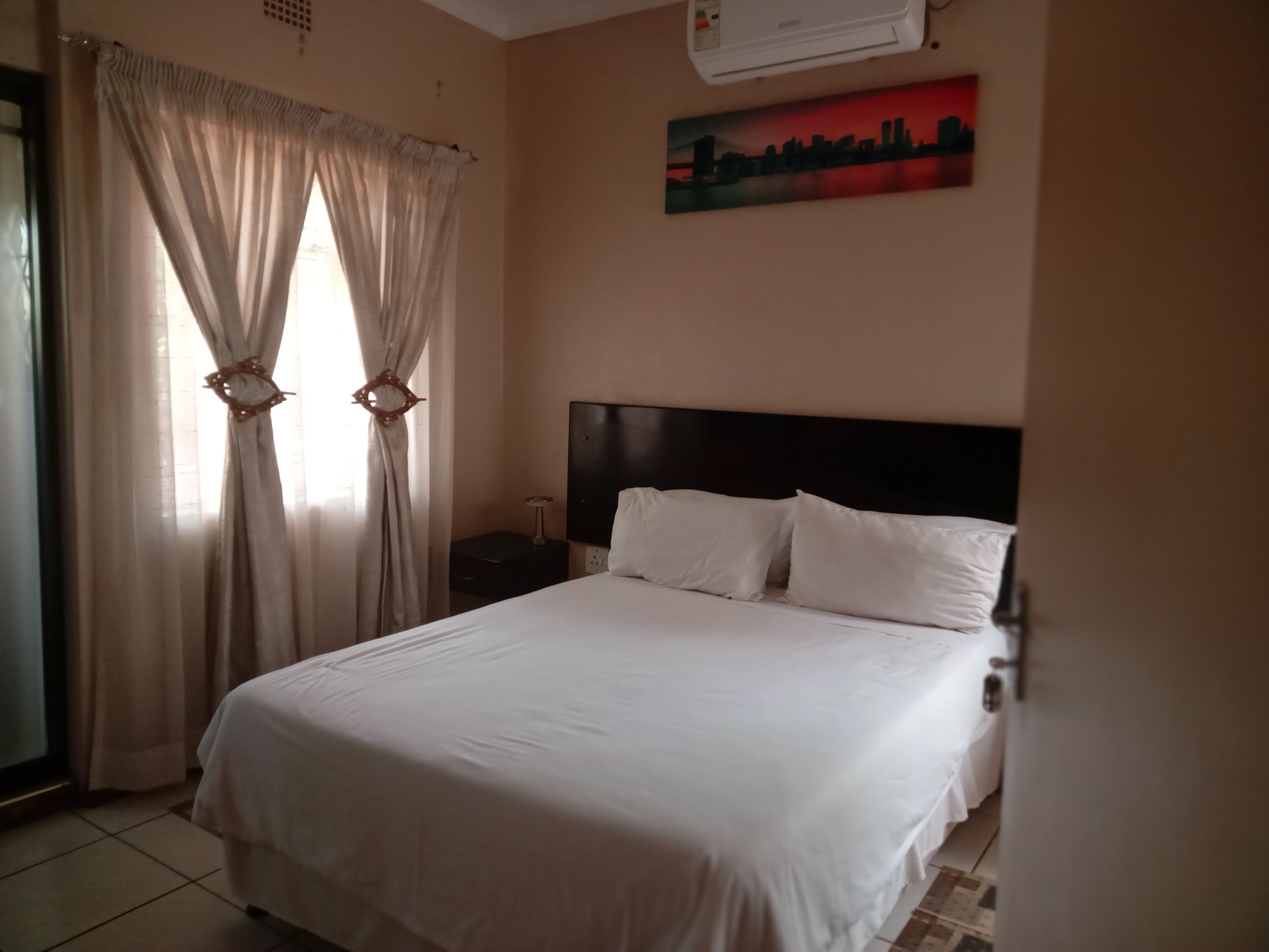 Habitación de hotel con cama, cortinas, mesita de noche, aire acondicionado instalado en la pared y una pintura de paisaje urbano.