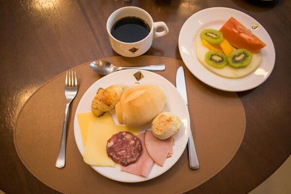 A breakfast setting with croissants, cheese, ham, sausage, and a plate of fruit including kiwi and tomato. - Hotel Jacques Georges Tower (Pelotas)