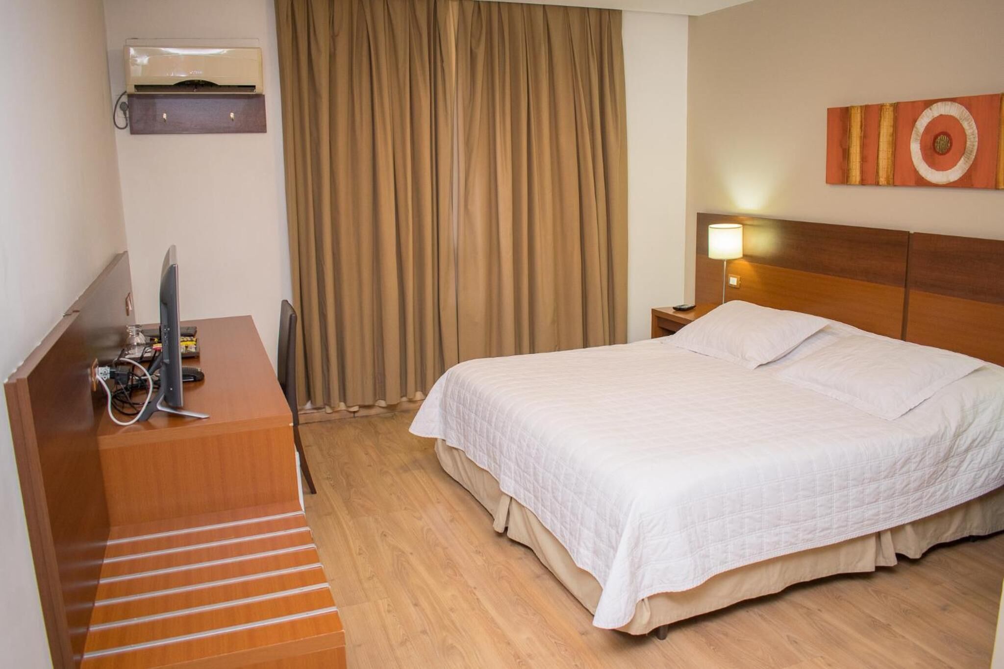 Quarto de hotel com cama, escrivaninha com computador, luminária e janela com cortinas.