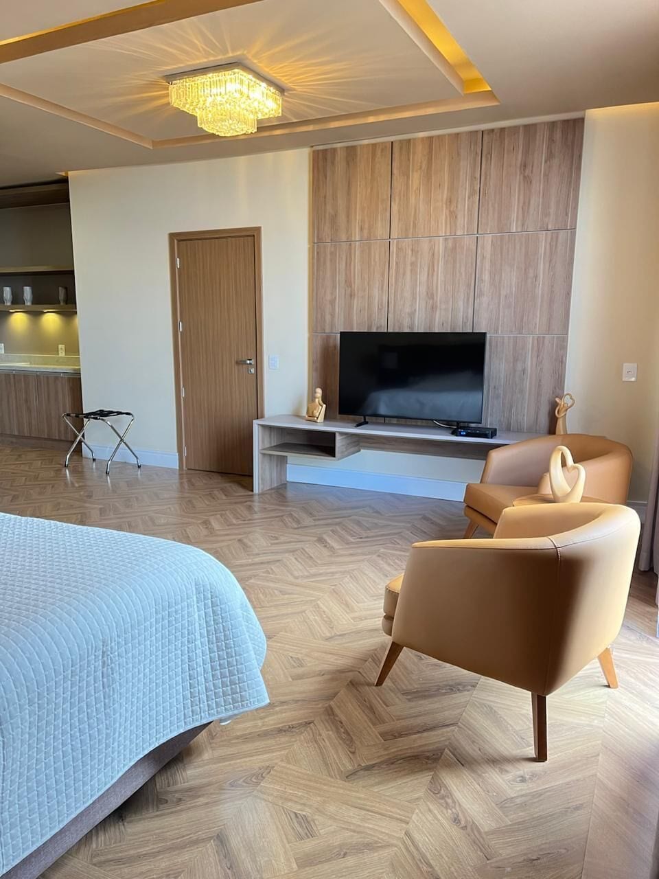 Una habitación de hotel moderna con una cama grande, un televisor montado en una pared de paneles de madera, una zona de estar con dos sillas y un suelo con estampado de espiga.