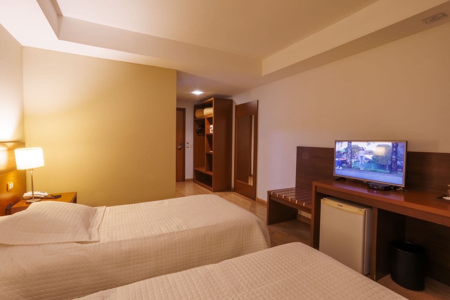 A hotel room with two beds, a television, a desk with a computer, and a small refrigerator.