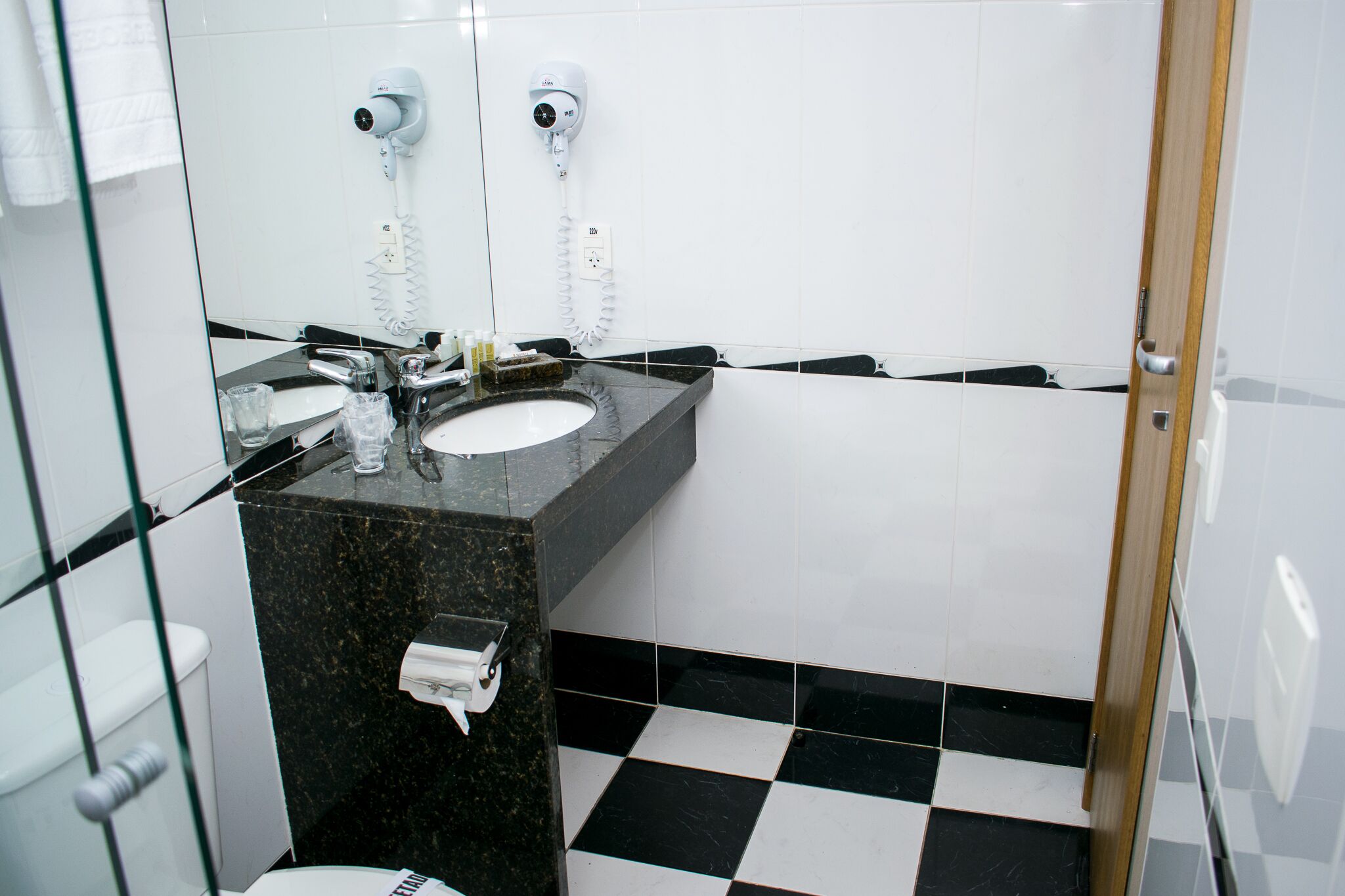 A bathroom with a black and white checkered floor, a sink with a marble countertop, and two hair dryers mounted on the wall.