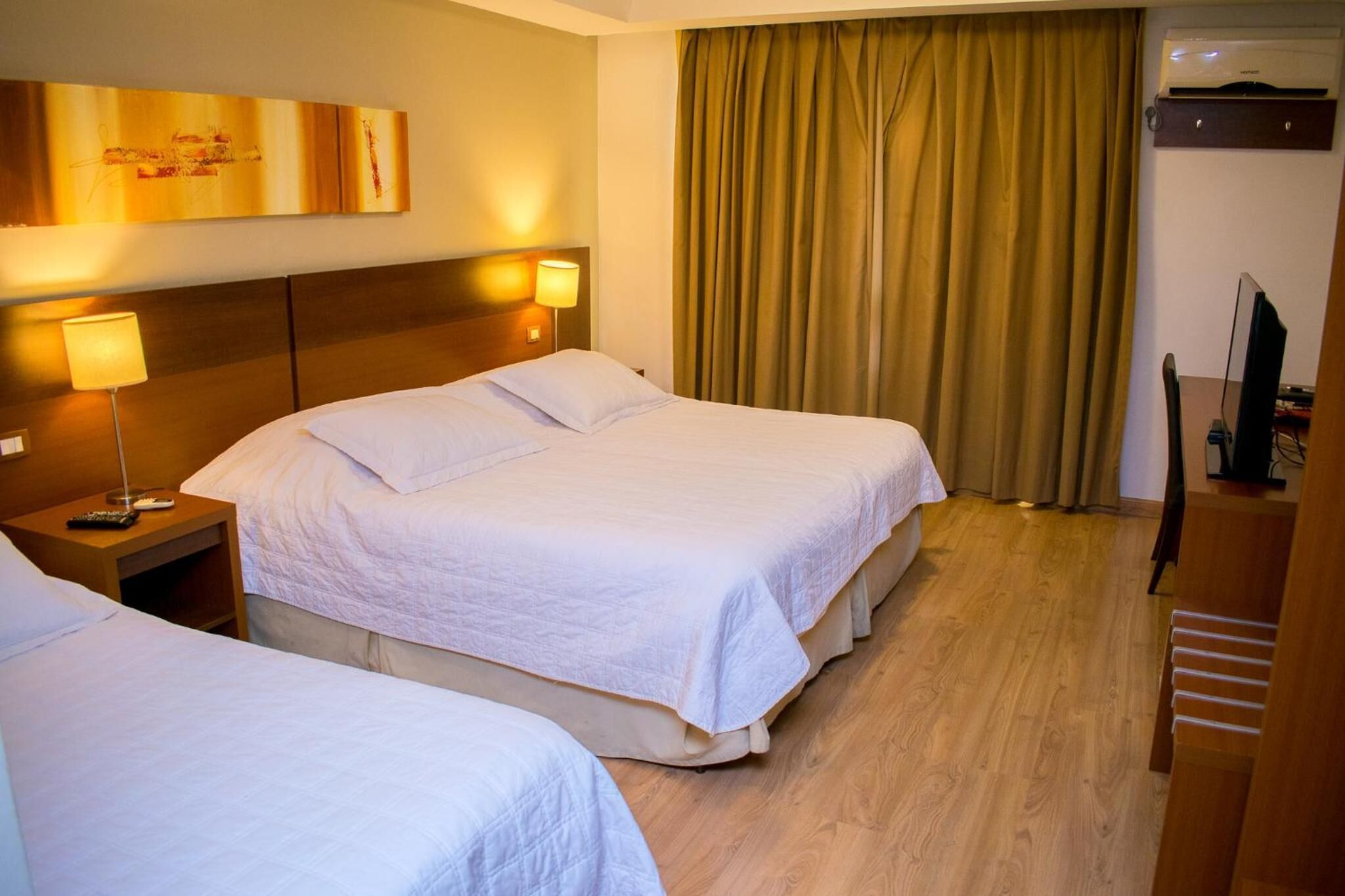 Une chambre d’hôtel avec deux lits, une tête de lit dotée d’une lampe, une grande fenêtre avec des rideaux et un téléviseur posé sur un meuble.