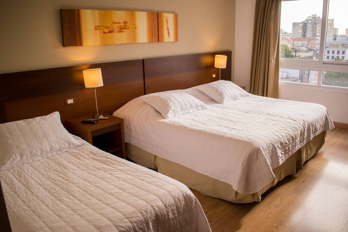 Habitación de hotel con dos camas, una mesita de noche con lámpara y ventana con vista a edificios.