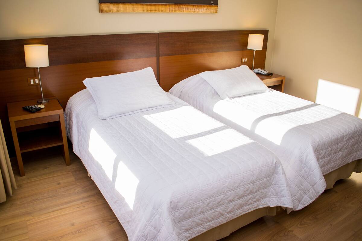 Una habitación de hotel con dos camas individuales, cabeceros de madera, lámparas de noche y suelo de madera.
