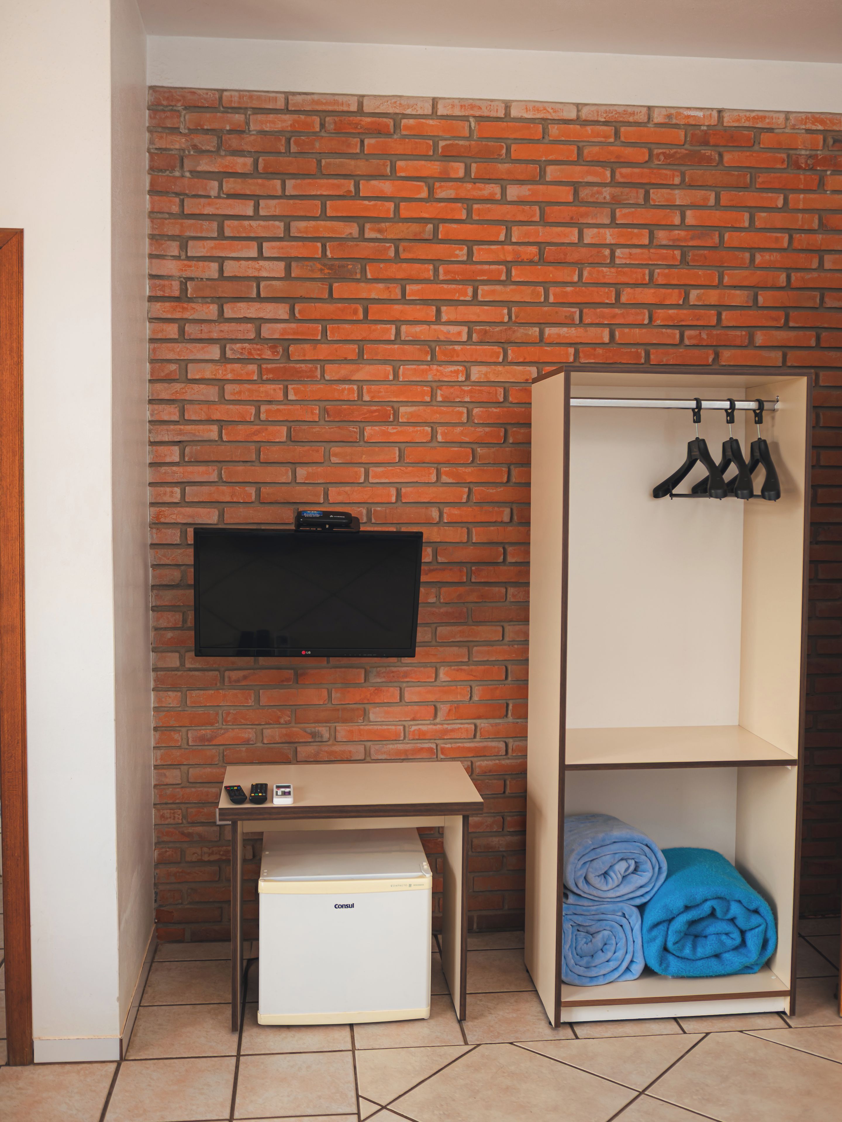 A room with a brick wall, a flat-screen TV mounted above a small desk, a wardrobe with hangers, and a rolled-up blue towel.