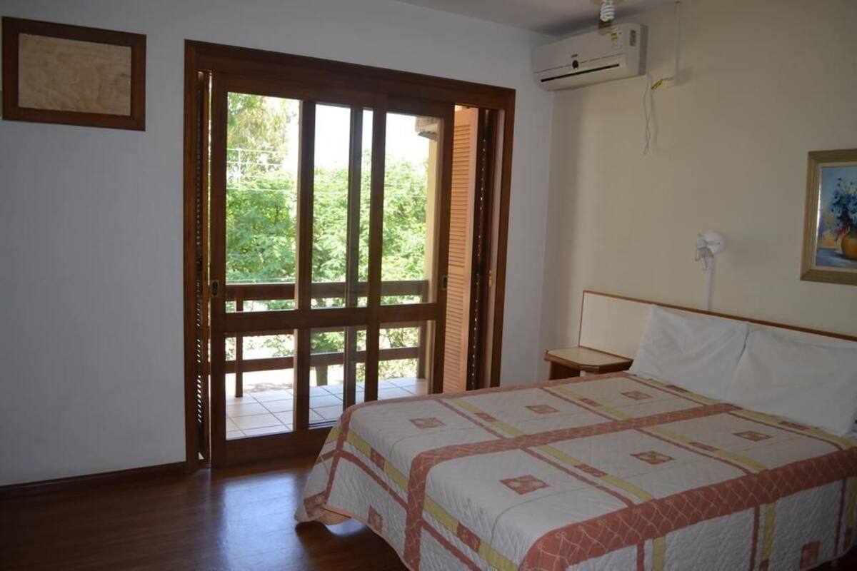 A bedroom with a bed, a nightstand, a window with a view of greenery, and an air conditioning unit.