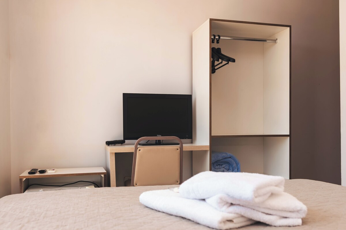 A hotel room with a bed, a desk with a television, a chair, and a wardrobe.