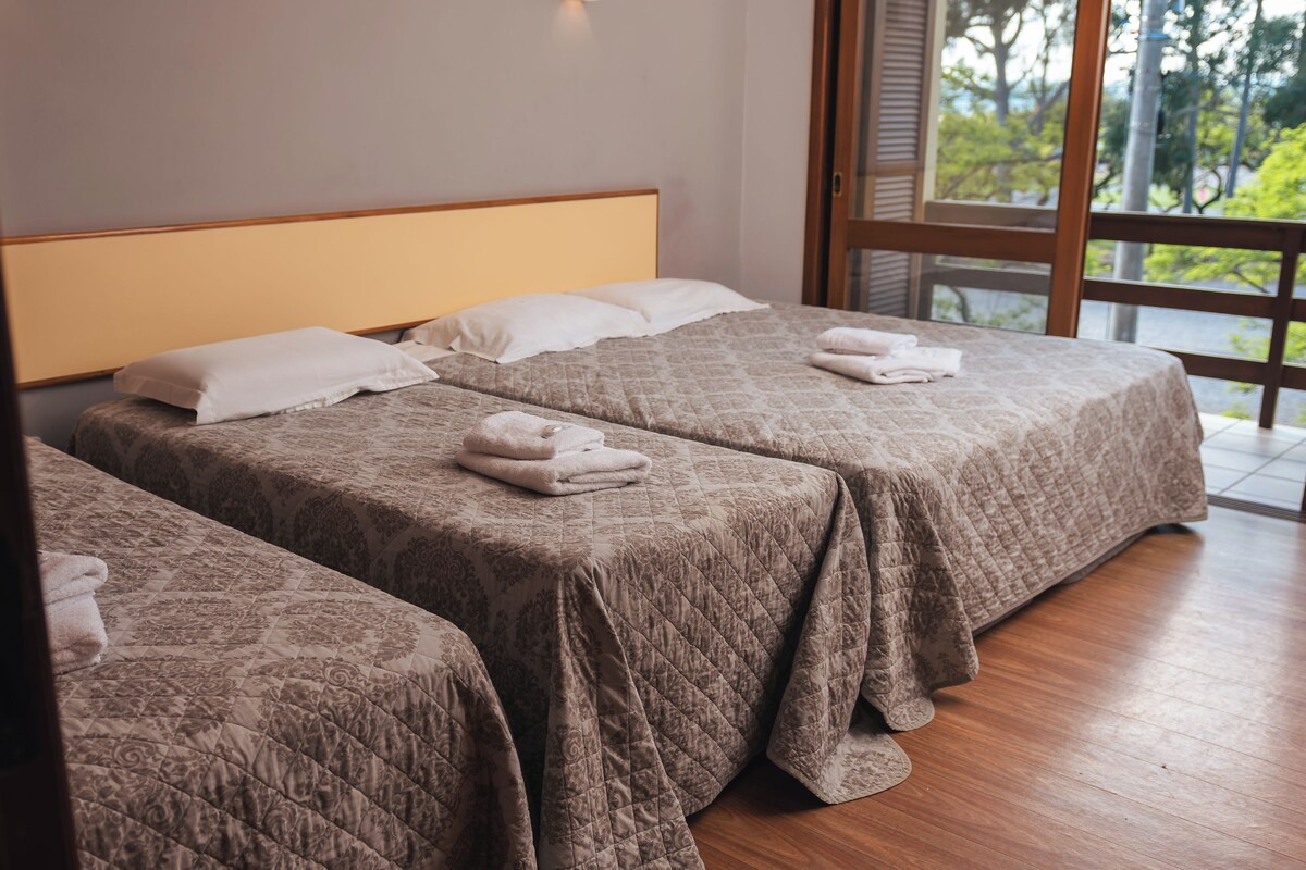 A hotel room with two beds, each with a quilted bedspread and pillows, and a large window offering a view of trees.