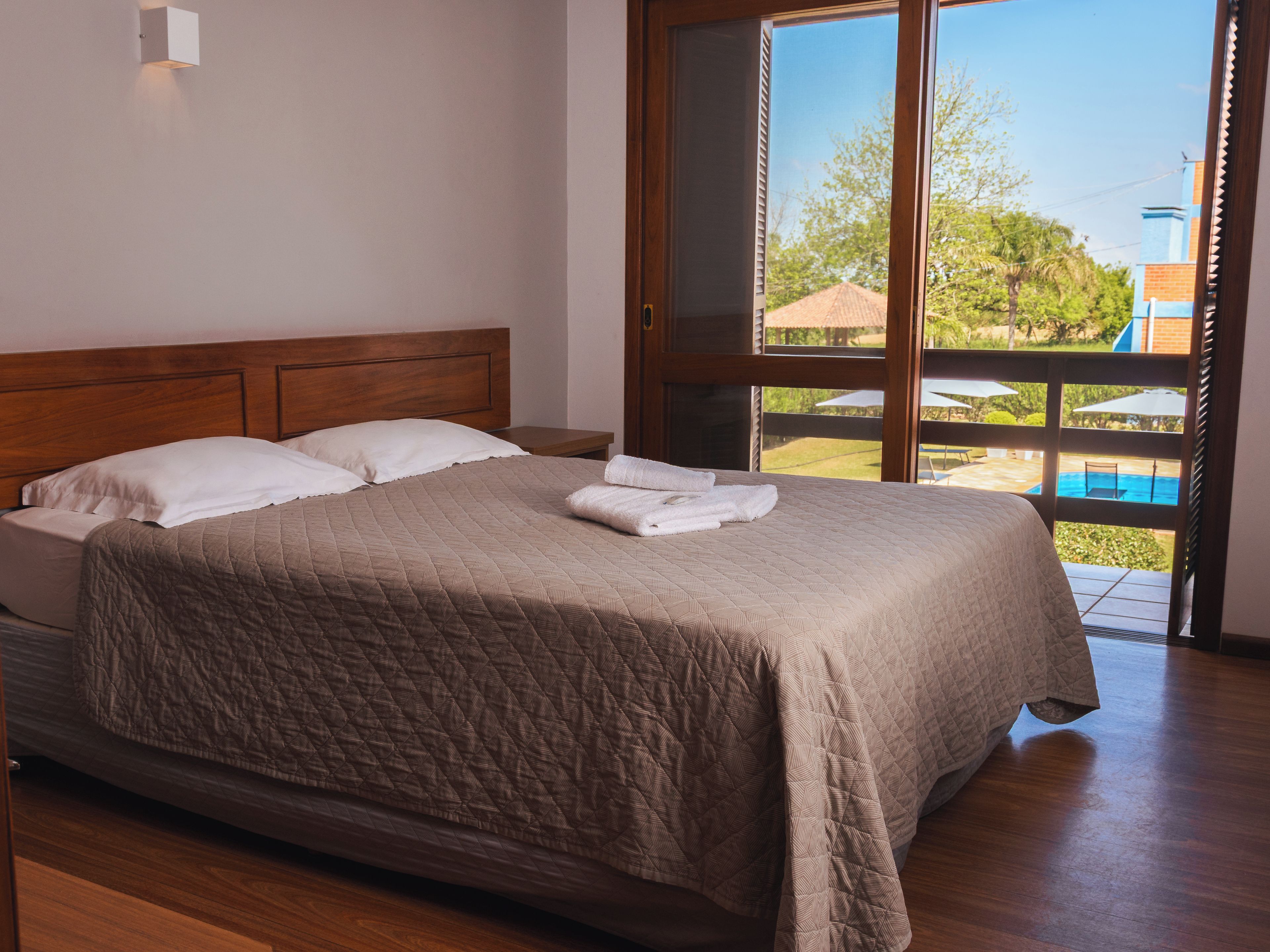 A bedroom with a large bed, wooden headboard, and a view of an outdoor swimming pool through a sliding glass door.