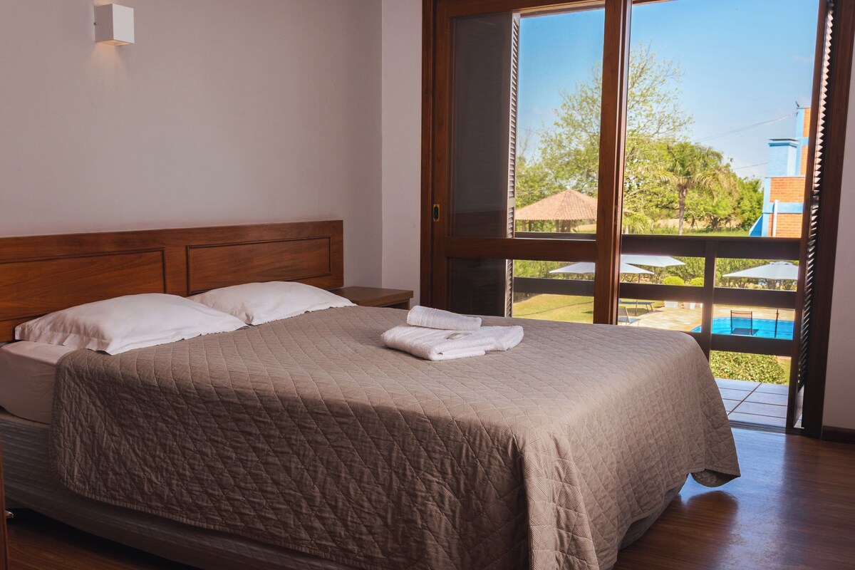 A bedroom with a large bed, wooden headboard, and a view of an outdoor swimming pool through a sliding glass door.