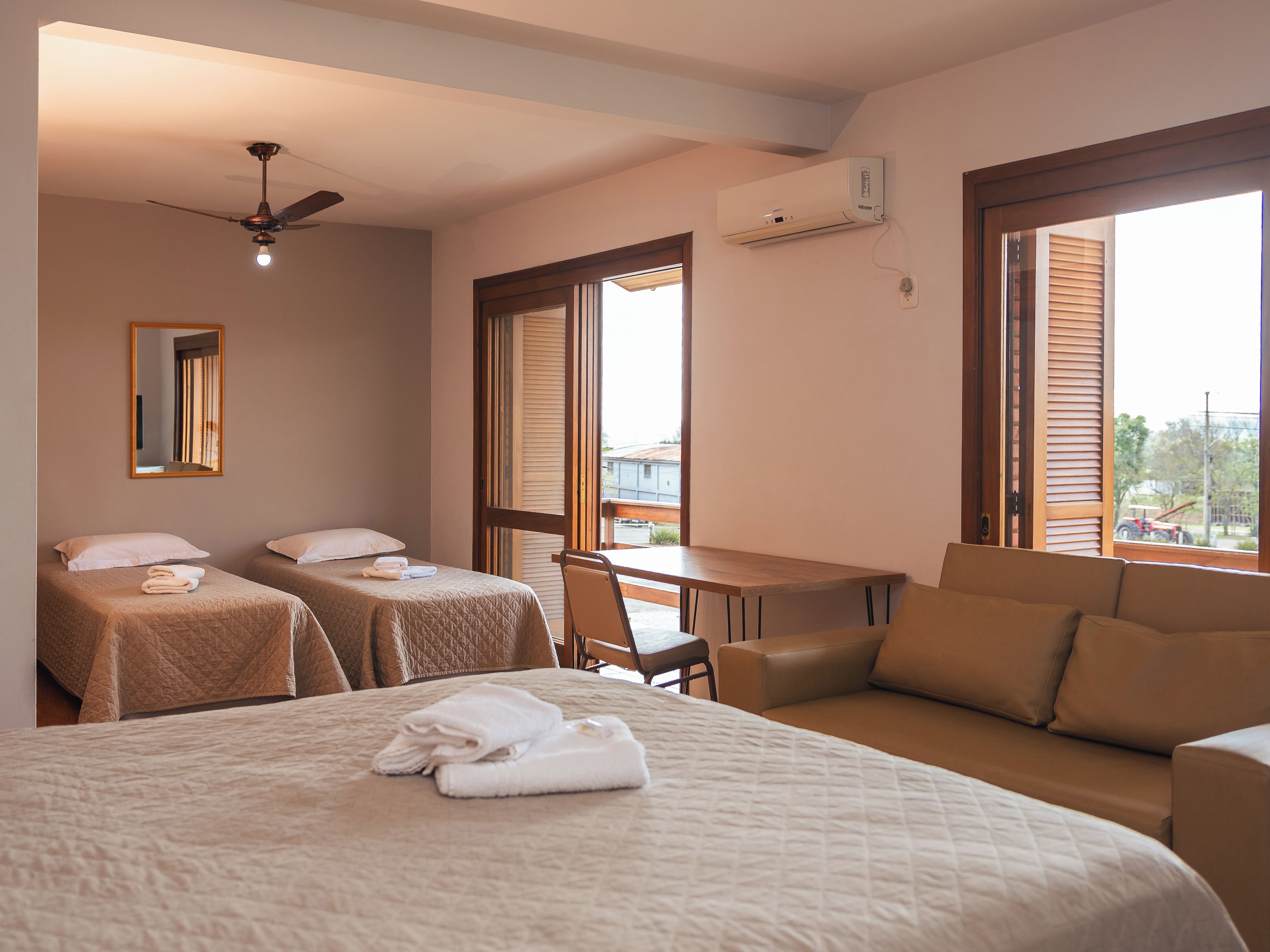 A hotel room with two beds, a desk, a chair, a ceiling fan, and an air conditioning unit.