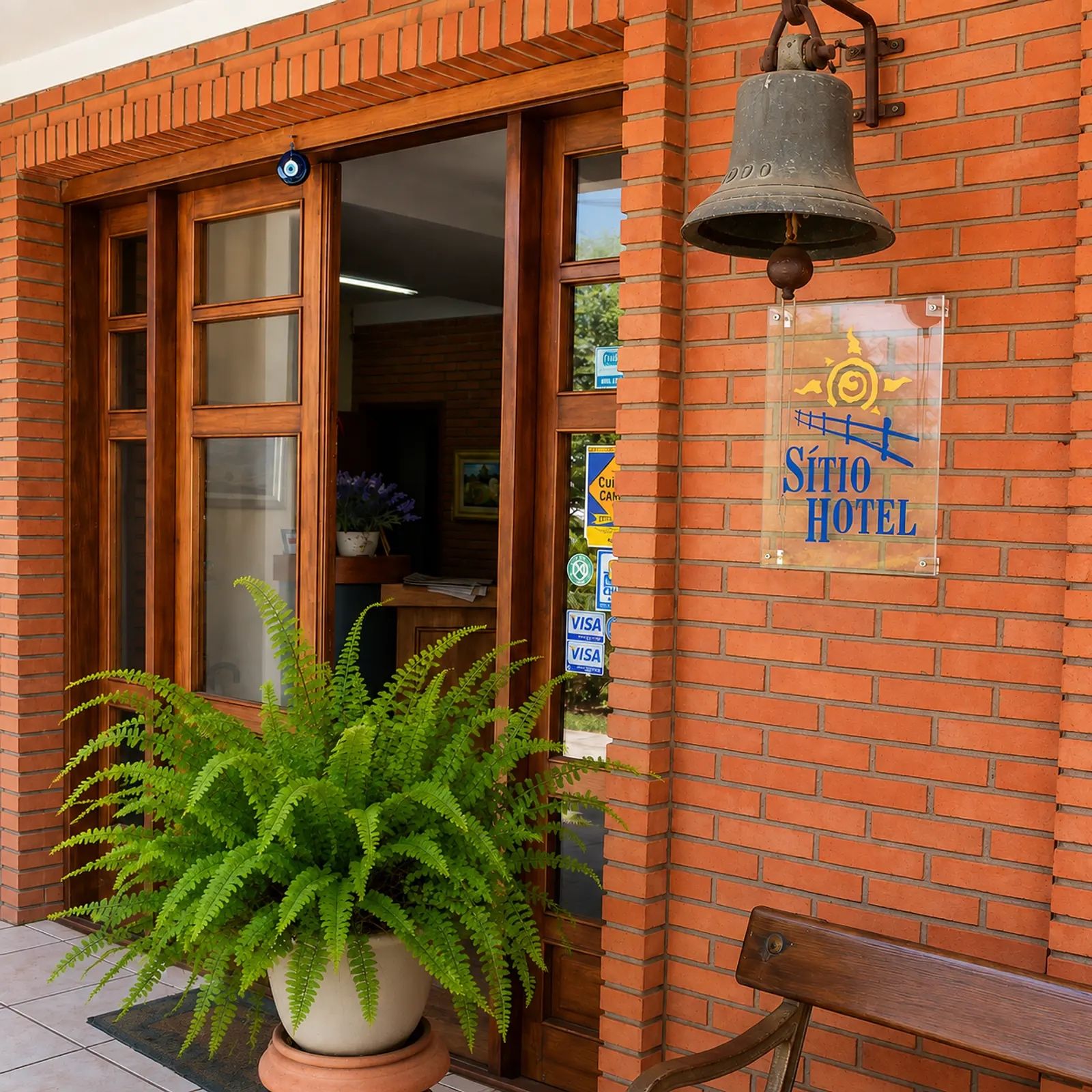 Entrada de un hotel con un helecho grande en maceta, un timbre y un letrero que dice Sìtio Hotel.