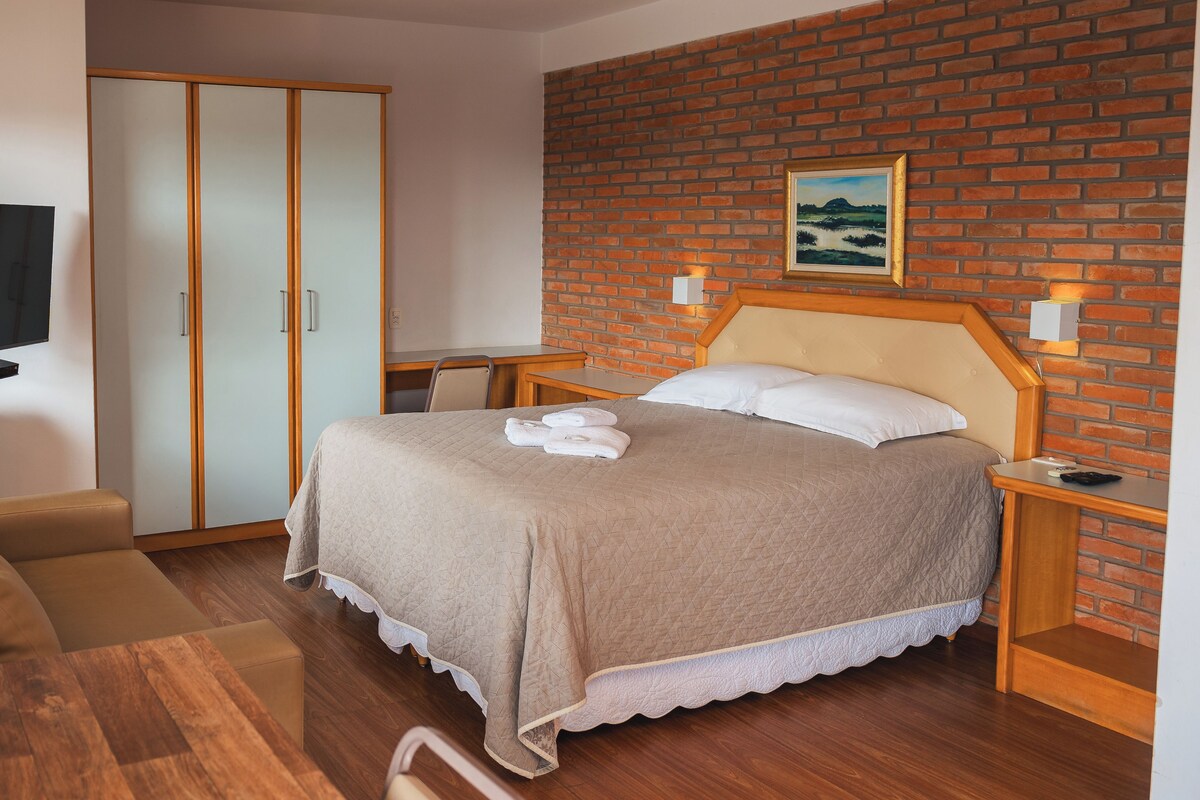 A bedroom with a bed, a wooden headboard, a brick wall, a painting, and a television on the wall.