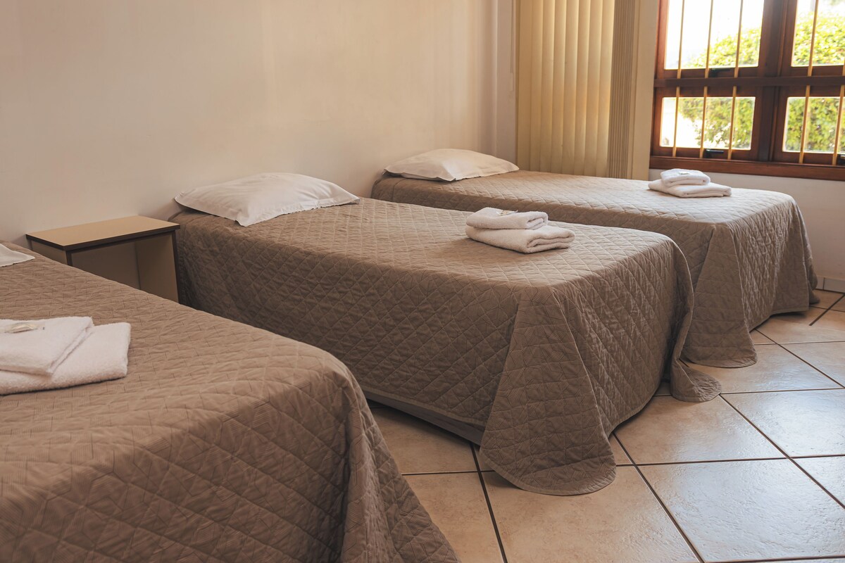 A hotel room with two beds, each with a quilted bedspread and a folded towel on top. There is a nightstand with a lamp, and the room has a window with blinds.