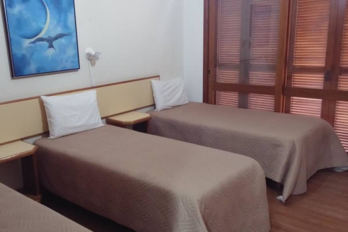A hotel room with two single beds, a nightstand, a painting of a bird and moon, and a window with blinds.