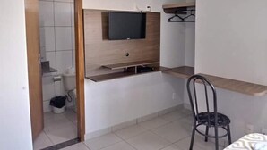 A compact room with a built-in TV, a small seating area, and a wooden shelf. - Hotel Guima (Santa Vitoria)