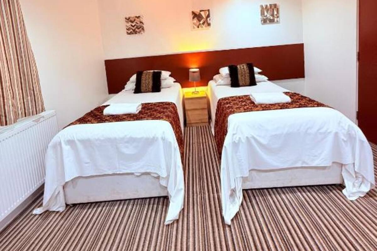 A hotel room with two beds, a radiator, a wooden headboard, a patterned carpet, and a wall-mounted artwork.