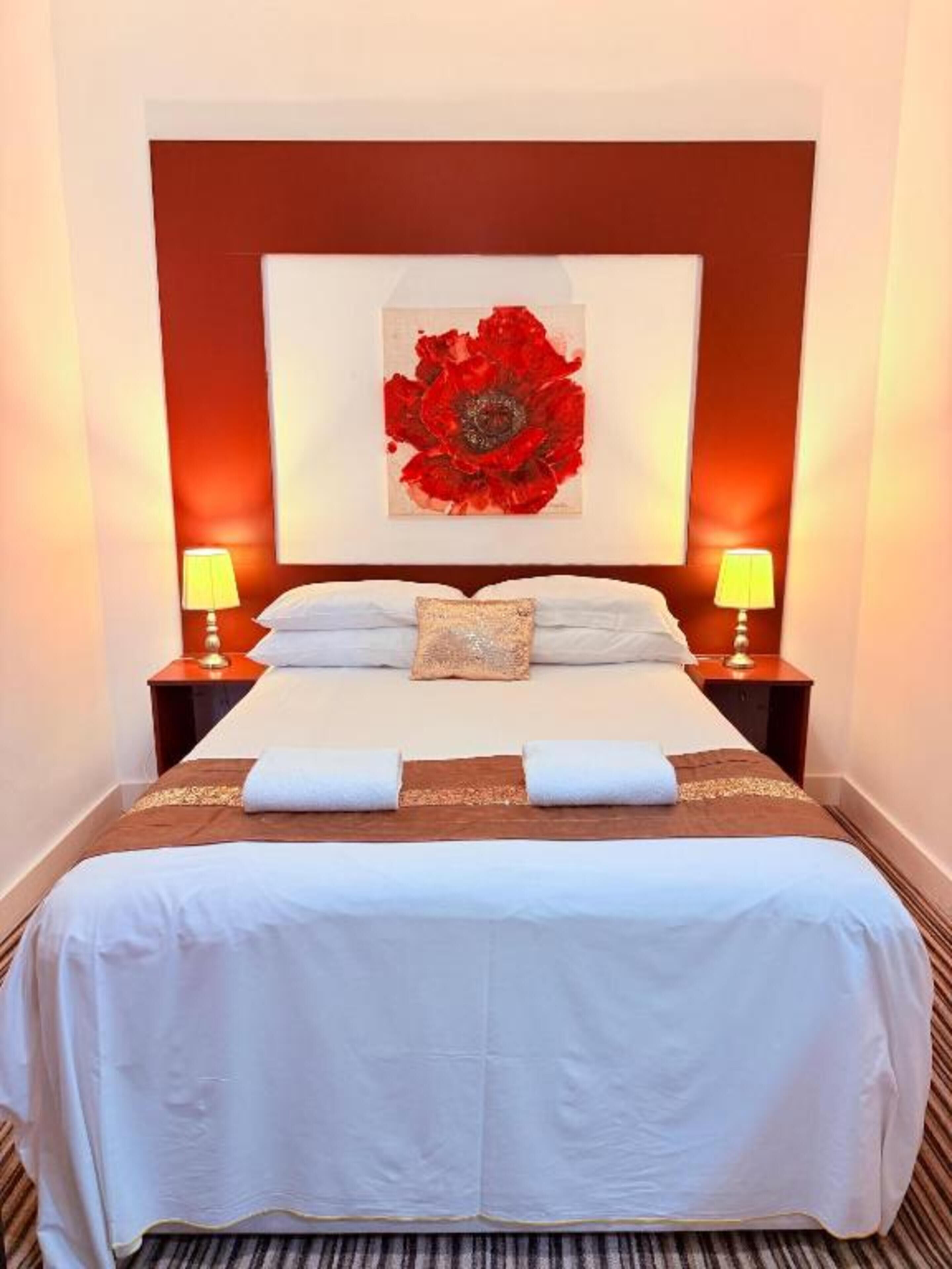 A hotel room with a bed, two bedside lamps, a large red floral artwork on the wall, and a neatly made bed with towels.