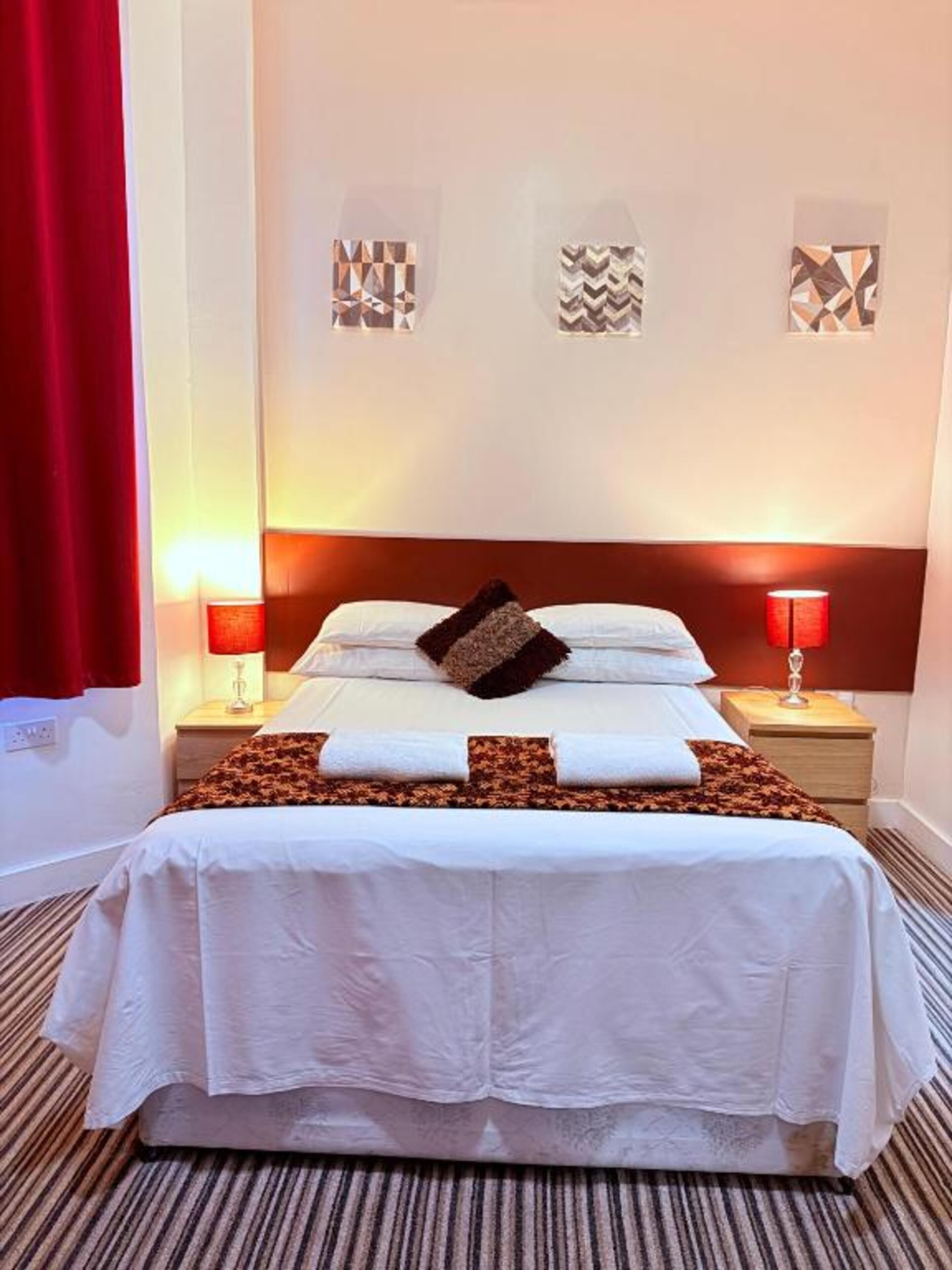 Une chambre d’hôtel avec un lit, deux tables de chevet avec des lampes, un rideau rouge et des motifs décoratifs sur les murs.