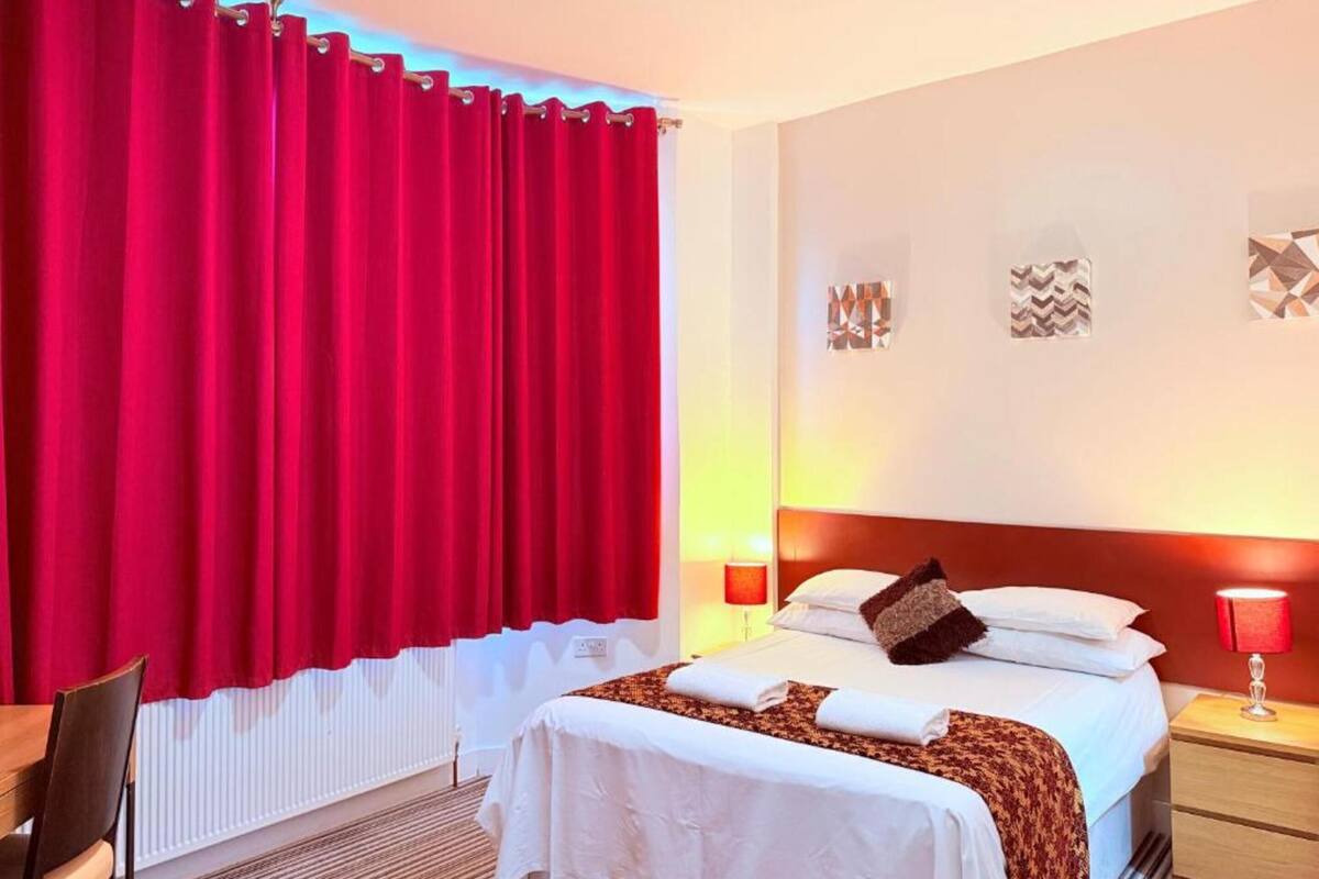 A hotel room with a bed, bedside tables, a chair, and a window with red curtains.