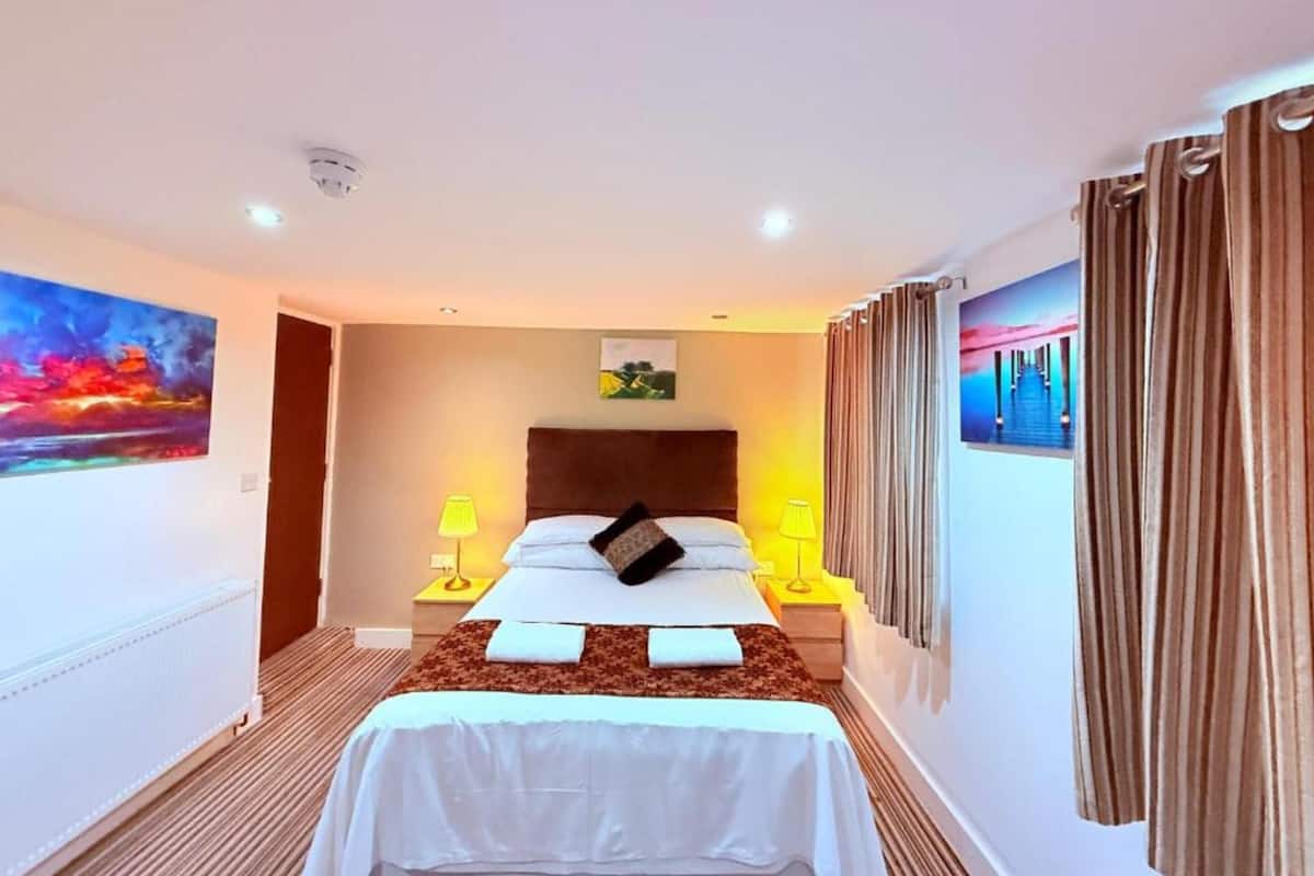 A hotel room with a bed, two bedside tables with lamps, a headboard, a painting on the wall, and a curtain.