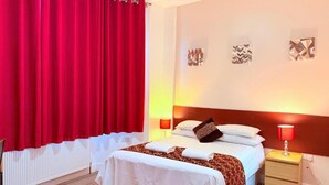 A hotel room with a bed, a nightstand, a lamp, and a window with red curtains. - RBS Hotel, Rochester (Rochester)