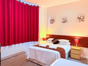 Una habitació d'hotel amb un llit, una tauleta de nit, una làmpada i una finestra amb cortines vermelles.
