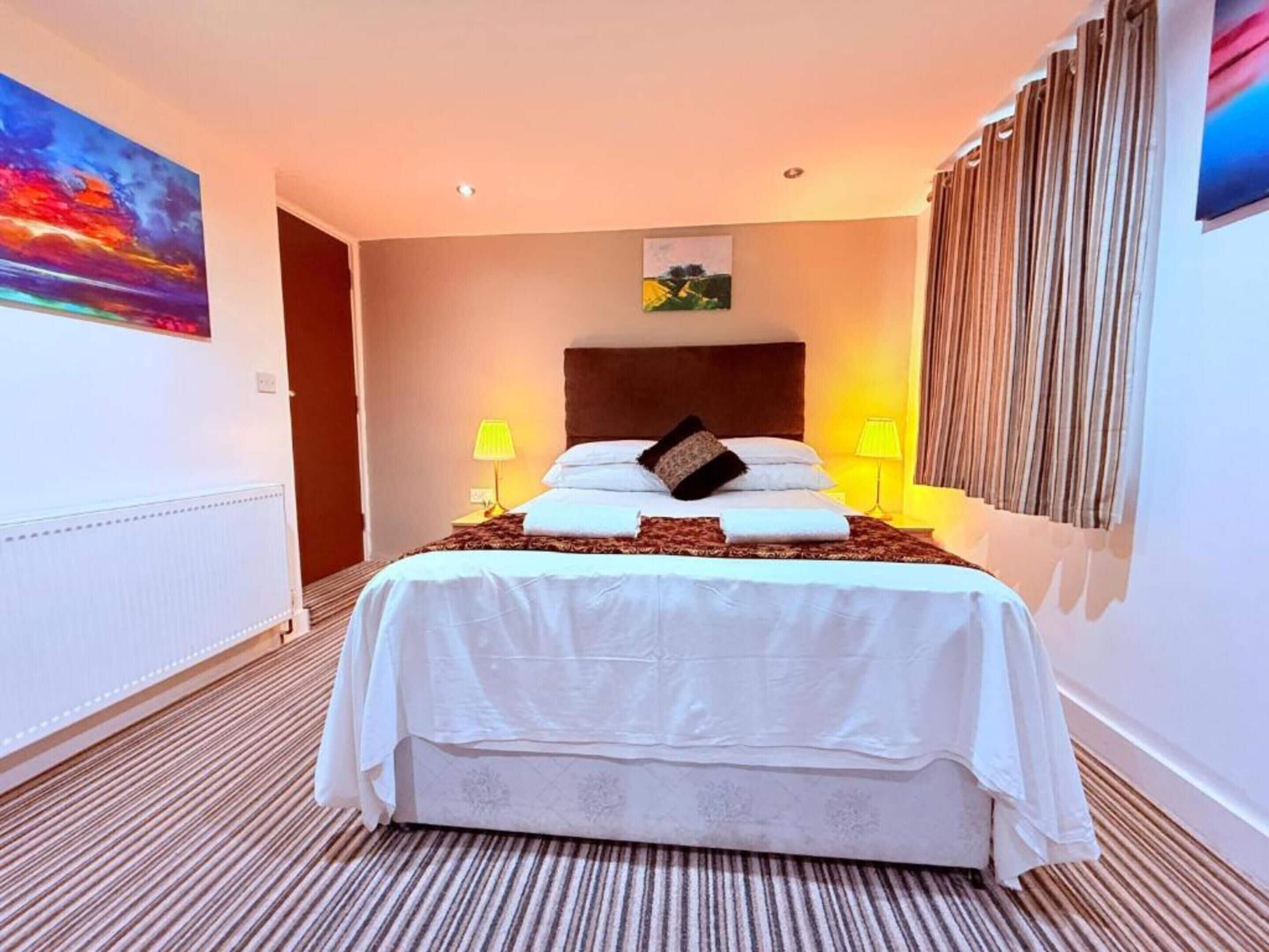 A hotel room with a large bed, two bedside lamps, a headboard, a painting on the wall, and a window with curtains.